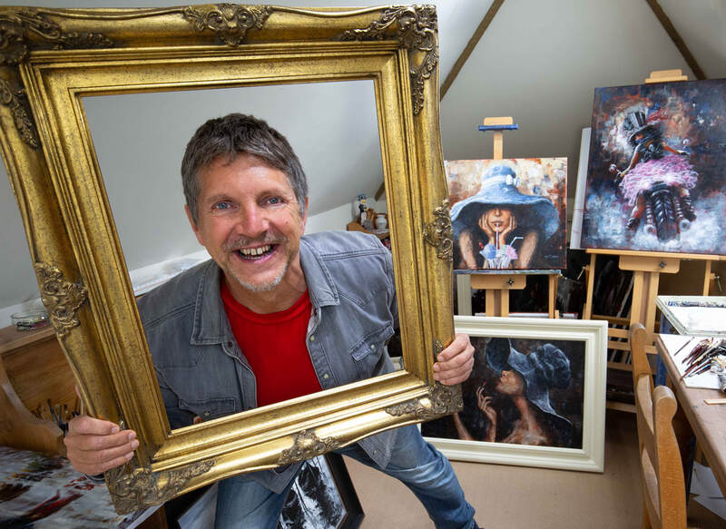 Neil Buchanan stands in a room full of paintings with his head in a picture fame.