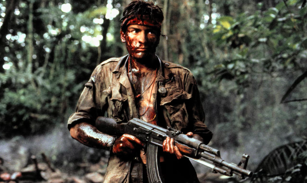 Charlie Sheen in Platoon wearing war-torn clothes and holding a large gun.